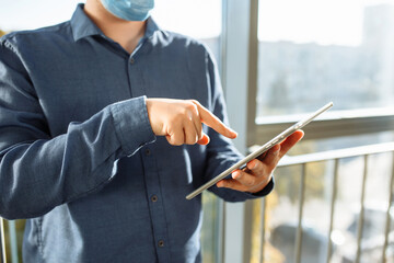 Young man with tablet in his hands standing at the office corridor wearing protective medical mask. Leading business and going to work during coronavirus pandemic. Stay safe concept.