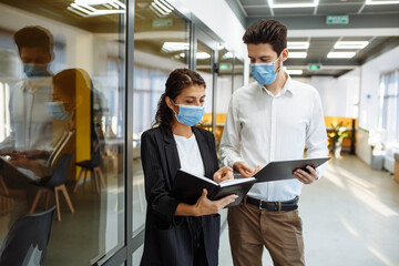 Businesspeople discuss workring issues during pandemic of coronavirus. Man and woman wearing protective medical masks talking business in the office corridor. Health safety at work concept.