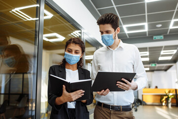 Businesspeople discuss workring issues during pandemic of coronavirus. Man and woman wearing protective medical masks talking business in the office corridor. Health safety at work concept.