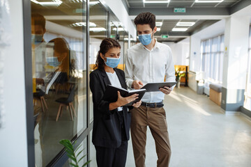 Businesspeople discuss workring issues during pandemic of coronavirus. Man and woman wearing protective medical masks talking business in the office corridor. Health safety at work concept.