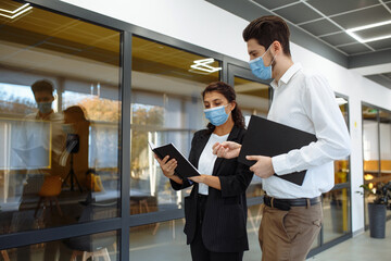Businesspeople discuss workring issues during pandemic of coronavirus. Man and woman wearing protective medical masks talking business in the office corridor. Health safety at work concept.