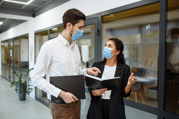 Businesspeople discuss workring issues during pandemic of coronavirus. Man and woman wearing protective medical masks talking business in the office corridor. Health safety at work concept.