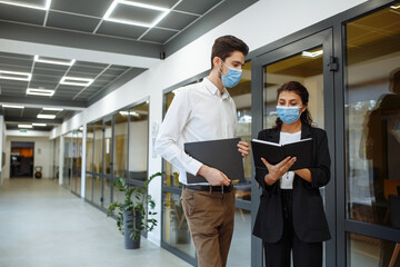Businesspeople discuss workring issues during pandemic of coronavirus. Man and woman wearing protective medical masks talking business in the office corridor. Health safety at work concept.