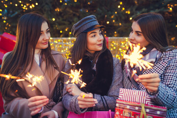 New Year concept. Women friends burning sparklers in Lviv by Christmas tree. Girls holding shopping bags and gifts