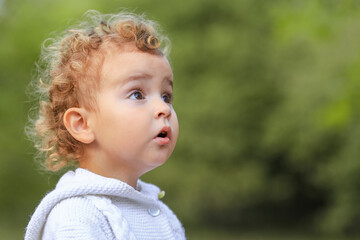 little curly surprised child in white knitted sweater, blurred green bokeh background, selective focus, place for text