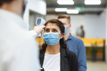 Office workers are checked with a distant non contact thermometer during coronavirus pandemic. Young business woman passing temperature control at the office corridor to prevent covid-19 spread.