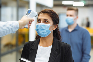Office workers are checked with a distant non contact thermometer during coronavirus pandemic. Young business woman passing temperature control at the office corridor to prevent covid-19 spread.