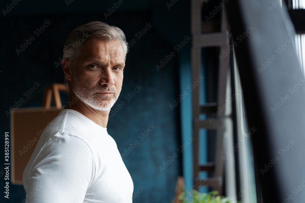 Wall mural smiling attractive casual grey-haired mature man standing near window looking at the camera