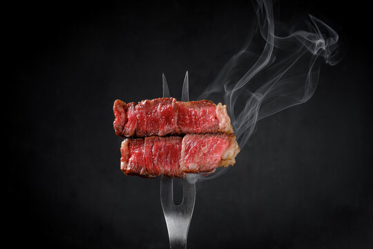 Grilled beef steak striploin on fork with smoke on dark background, close up