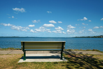 Fototapeta na wymiar A place to sit and relax. Bench seat beside the water, with a view across the bay to islands on the horizon, and blue sky with fluffy clouds. Victoria Pt, Moreton Bay, Redlands, Queensland, Australia.