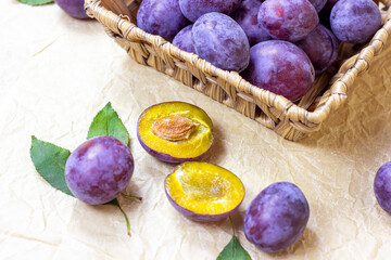 Fresh raw blue and violet plums in wooden bowl with green leaves on light paper background.