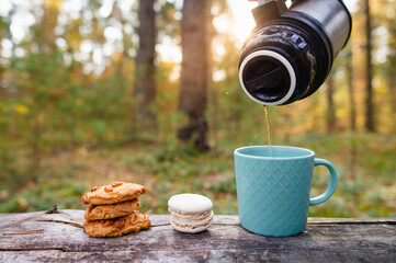 A cookie and a mug are on a log in the forest. Hot tea is poured from a thermos into a mug. Picnic in the autumn forest.