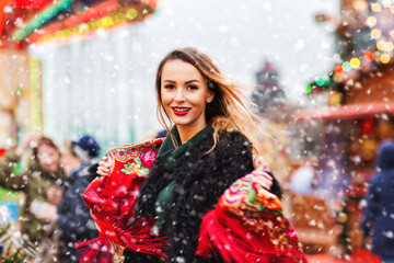 Outdoors lifestyle portrait of pretty blonde young woman. Smiling and dancing on the christmas market. Wearing stylish black fur coat, shawl with a floral pattern in the Russian style. Festive mood