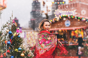 Outdoors lifestyle portrait of pretty blonde young woman. Smiling and walking on the christmas market. Wearing stylish shawl with a floral pattern in the Russian style. It's snowing. Festive mood