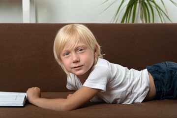 Blond schoolboy is lying on couch with book. Portrait of boy with textbook