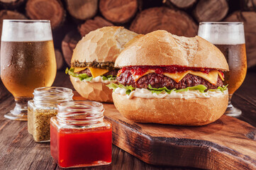 Two burger with pepper jelly, pesto sauce and beer with firewood pile in background