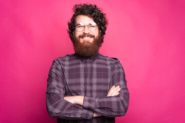 A nice young man with a beard is standing with his arms crossed near a pink wall with glasses on