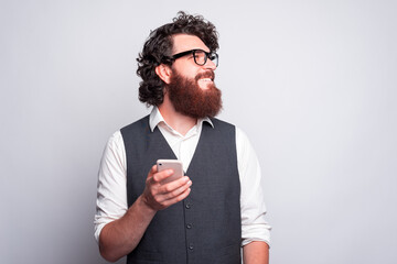 Young bearded man is holding a phone looking away is smiling with his glasses on