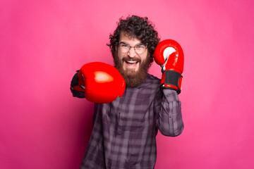 Young bearded and cheerful man with boxing gloves on and glasses is looking at the camera