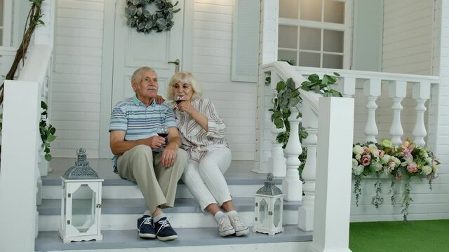 Attractive senior elderly Caucasian couple sitting and drinking wine in porch at home, making a kiss