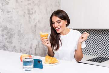 Young woman sitting at the kitchen look video on mobile phone while eat breakfast croissant in the morning