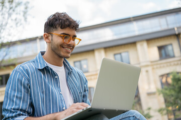 Portrait of happy Indian programmer using laptop computer, internet, working freelance project online, sitting in park. Asian student studying, learning language, using modern technologies outdoors
