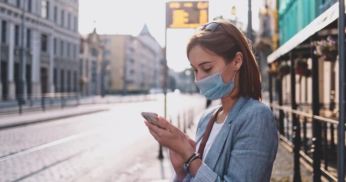 Young Woman In Protective Mask Using SmartPhone, Waiting For Tram In Sunny Morning City. SLOW MOTION, Gimbal Stabilizer. BusinessWoman in face mask against air pollution and coronavirus use transport.