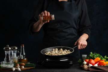 The professional chef in black uniform pours spices in pan with meat. Backstage of preparing grilled meat for dinner on dark blue background. Food concept. Frozen motion. Traditional recipe.