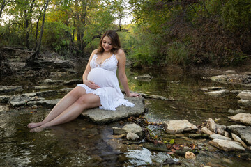 Pregnant expecting young mother sitting on rock on the water creek looking down at maternity belly outdoor portrait 