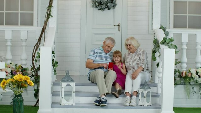 Senior ouple sitting with granddaughter in porch at home. Using mobile phone for online video call