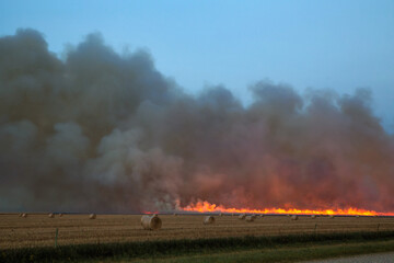 Fototapeta na wymiar A line of fire with billowing smoke behind a field dotted with straw bales in an autumn evening landscape