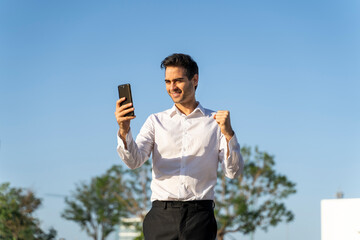 Happy businessman gesturing while using smart phone against clear blue sky