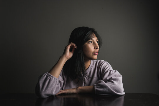 Thoughtful woman with black hair sitting at table against wall