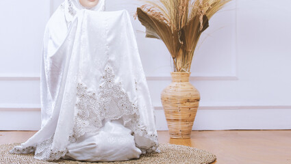 A Muslim woman wearing moslem clothes with hijab called mukena, praying by raising her hands with minimalist home background 