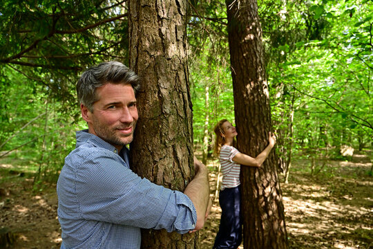 Smiling man and woman hugging tree while standing in forest