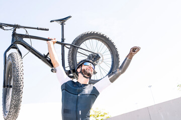 Male amputee cyclist with arms raised carrying bicycle while standing against clear sky