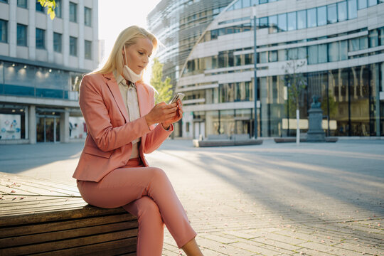 Female professional using smart phone while sitting on bench at financial during coronavirus