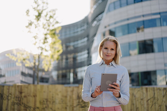 Confident businesswoman holding digital tablet while standing against building at financial district