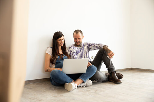 Couple using laptop while sitting against white wall in new unfurnished home
