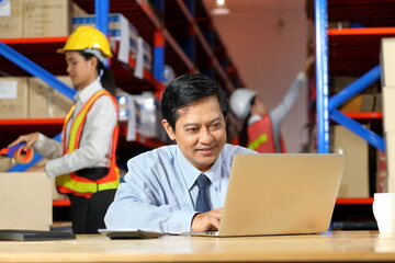 Asian warehouse manager is working with laptop computer while employees are checking the inventory for industrial and stock merchandise concept