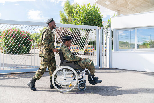 Army soldier helping military soldier on wheelchair while standing against gate on sunny day