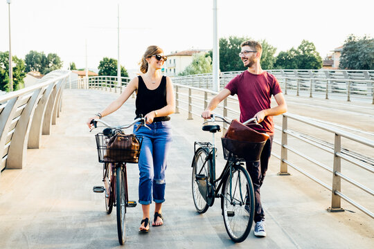 Happy couple talking while walking with bicycles on bridge against clear sky