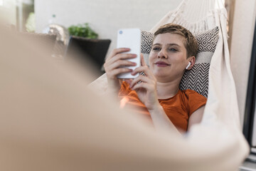 Mid adult woman with wireless headphones using smart phone while lying on hammock in porch