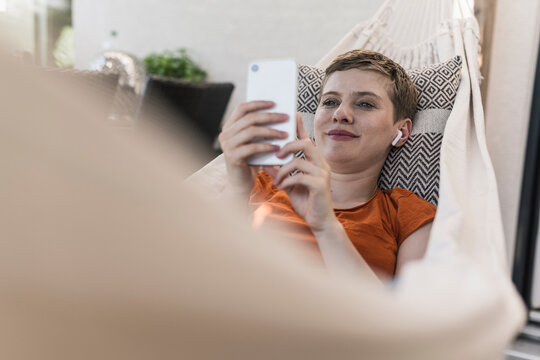 Mid adult woman with wireless headphones using smart phone while lying on hammock in porch