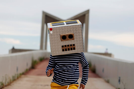 Boy wearing robot mask made of box running on footpath against sky