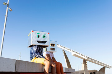 Boys wearing robot masks made of boxes while sitting on retaining wall against clear blue sky