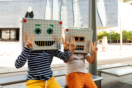 Boys wearing robot masks showing peace sign while sitting on seat at railroad station