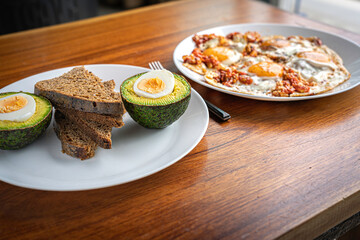 Close up fresh avocado cut in half, slices of bread and fried eggs with tomato on white plates on wooden table