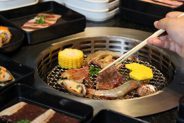 Grilled pork meat and seafood in chopsticks on hot steam smoke water from hot pan for cooking in party, roasted meat on night background for people eating to tasty and delicious food in restaurant