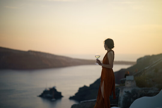 Woman tourist in gorgeous dress with glass of wine relaxing at sunset view at whitewashed village in Oia, Santorini Greece. mediterranean sea.